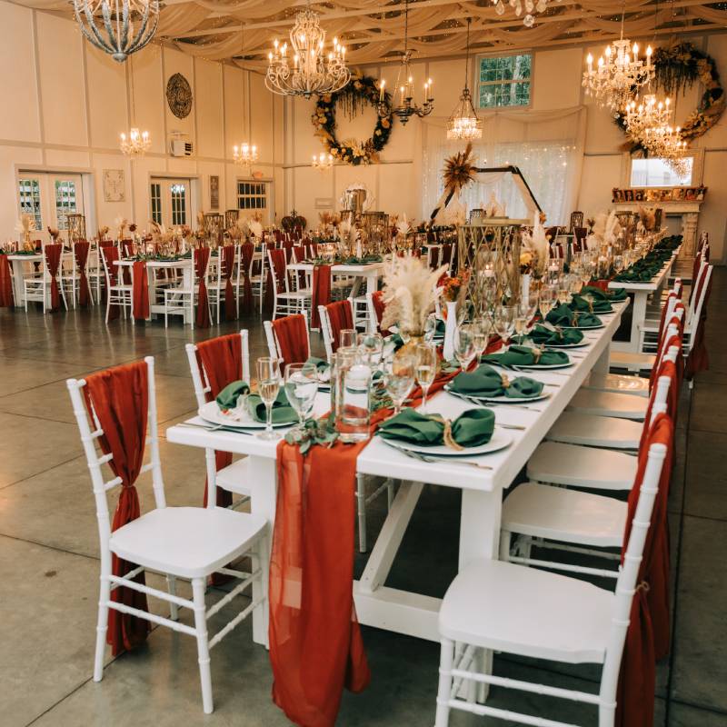 Long white farm tables inside the Manor Barn, set with sage napkins and rust chair sashes beneath crystal chandeliers