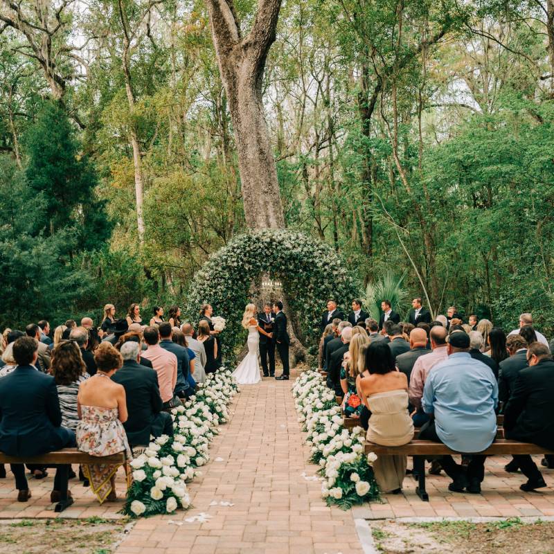 Outdoor ceremony beneath the live oak at Saxon Manor, with a white floral arch and a rose-lined brick aisle