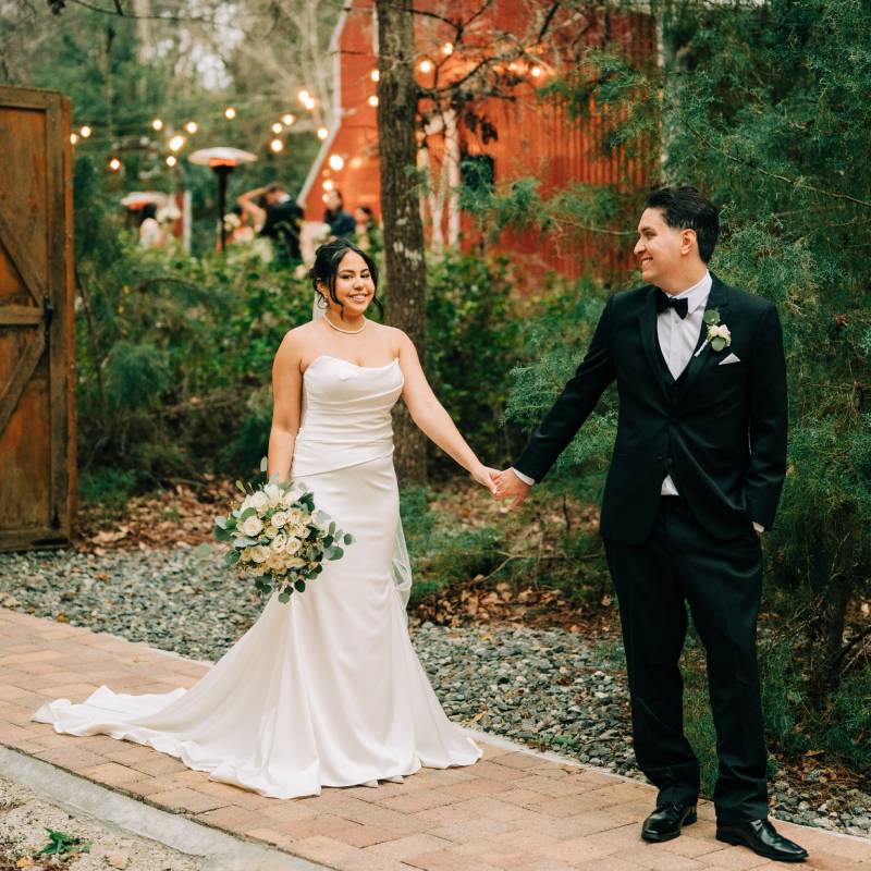 Bride and groom holding hands on the brick path outside the red barn, string lights overhead