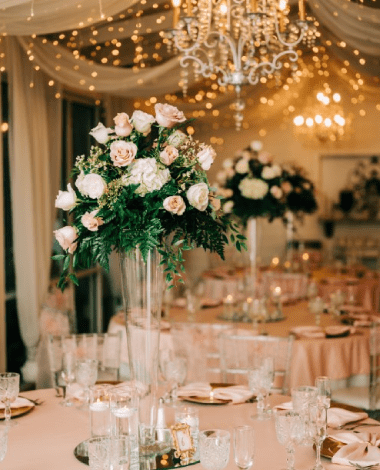 A tall rose centerpiece on a blush-linen table in the Garden Room, under chandeliers and string lights