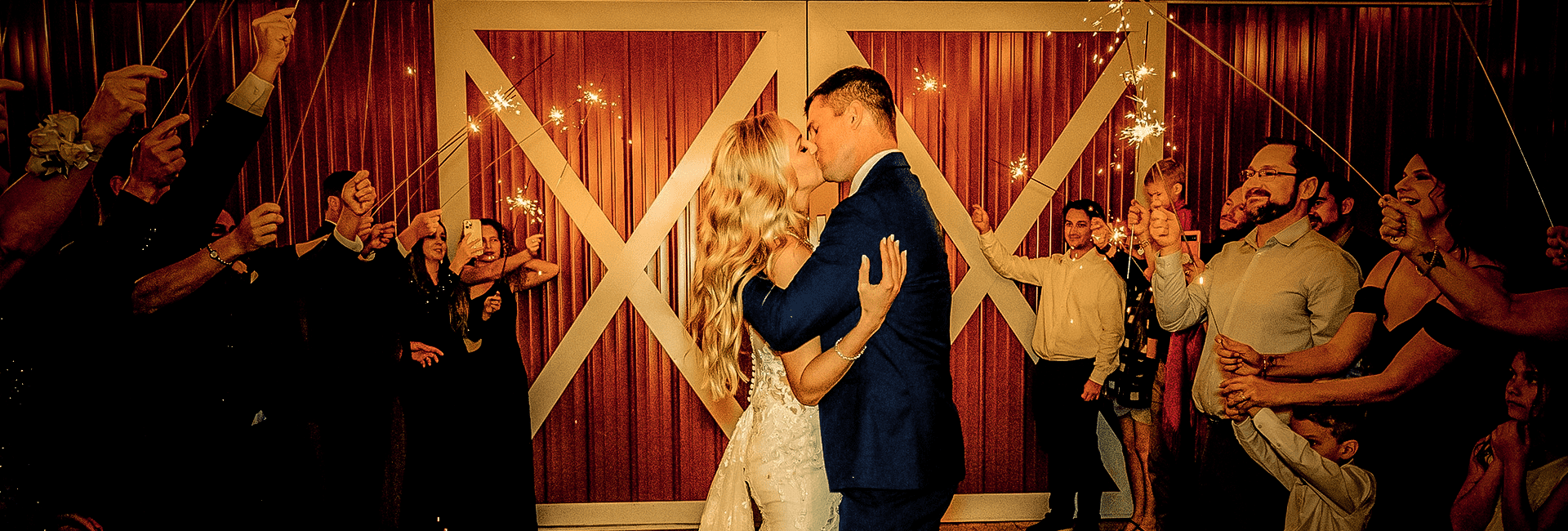 Sparkler send-off at night, the couple kissing in front of the barn’s white X-braced doors