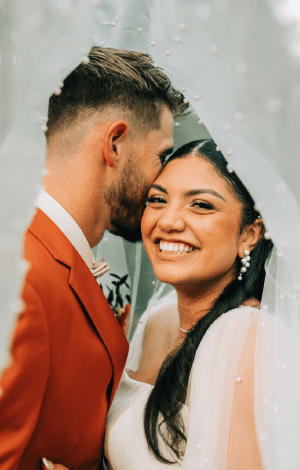 Bride and groom close together beneath her veil, the groom in a rust suit