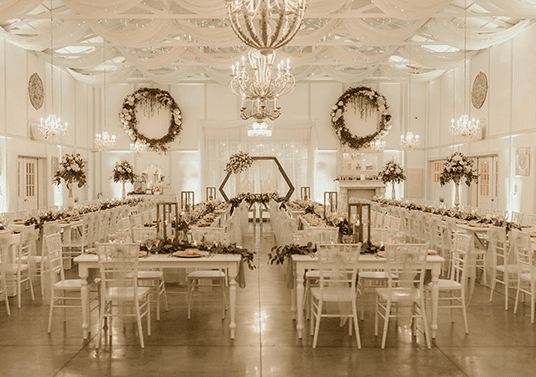 The Manor Barn set for dinner — long white tables and chiavari chairs under a draped ceiling and chandeliers