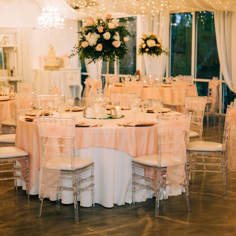 Round tables in the Garden Room dressed in blush linens with tall rose centerpieces, glass walls open to the gardens