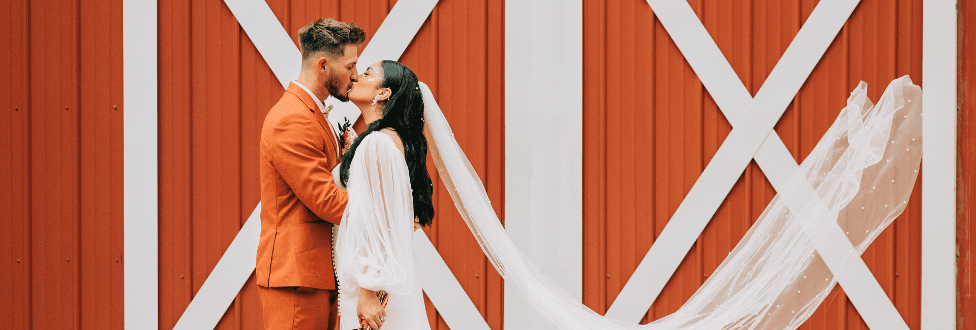 Bride and groom kissing against the red barn doors at Saxon Manor, the bride's veil caught in the wind